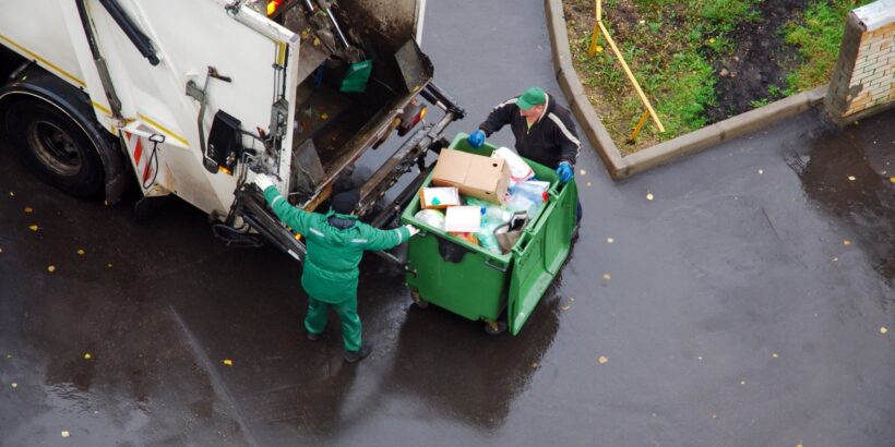A garbage truck picks up a business's trash from their large green hopper outside. There are two garbagemen sorting.