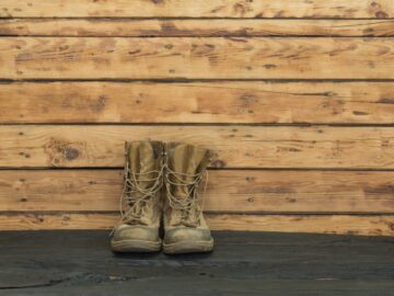 A pair of tan-colored military boots sit up against a wooden wall. They stand on a blacktop ground.