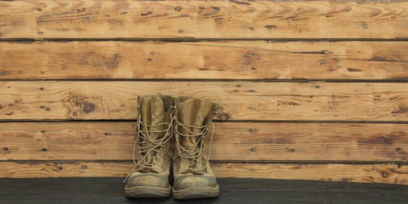 A pair of tan-colored military boots sit up against a wooden wall. They stand on a blacktop ground.