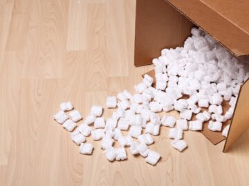 An overhead view of an open cardboard box on a hardwood floor with white packing peanuts spilling out.