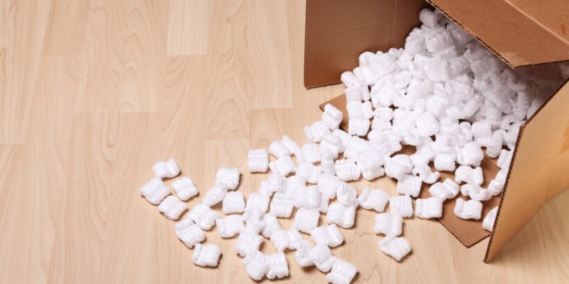 An overhead view of an open cardboard box on a hardwood floor with white packing peanuts spilling out.
