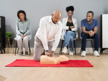A Black man wearing a white collared shirt performs CPR on a dummy in front of three other people sitting behind him.