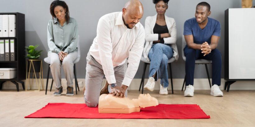 A Black man wearing a white collared shirt performs CPR on a dummy in front of three other people sitting behind him.