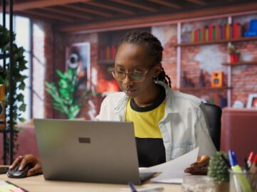 A young person sitting a desk in their at-home remote workspace. They are intently using a silver laptop.