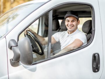 A man wearing a white polo shirt and a black cap, sitting inside a white car behind the wheel while smiling.