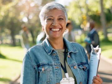 A smiling woman with short grey hair is wearing a black lanyard around her neck and holding a clipboard in a sunny outdoor space.