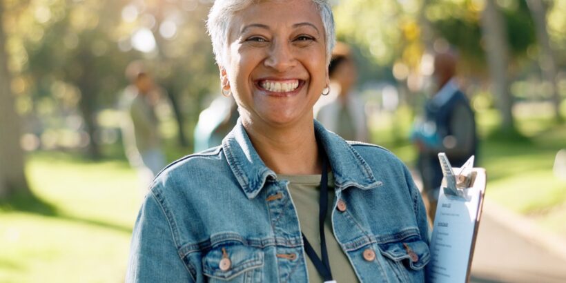 A smiling woman with short grey hair is wearing a black lanyard around her neck and holding a clipboard in a sunny outdoor space.