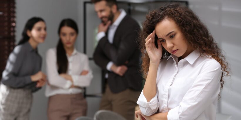An upset woman standing apart from her coworkers. Behind her, three coworkers are staring at her and talking.