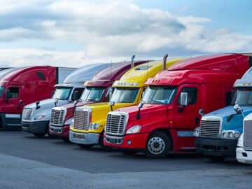 Multiple red, yellow, and white semi-trailer trucks line up in a row, parked on asphalt, underneath a cloudy sky.