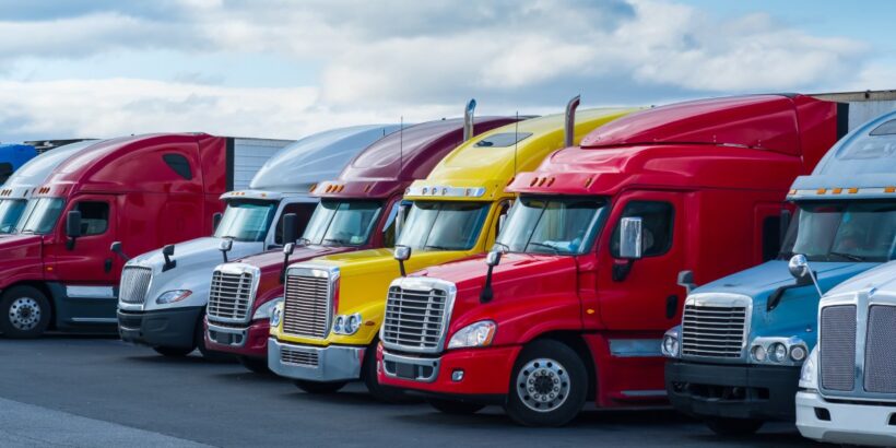 Multiple red, yellow, and white semi-trailer trucks line up in a row, parked on asphalt, underneath a cloudy sky.