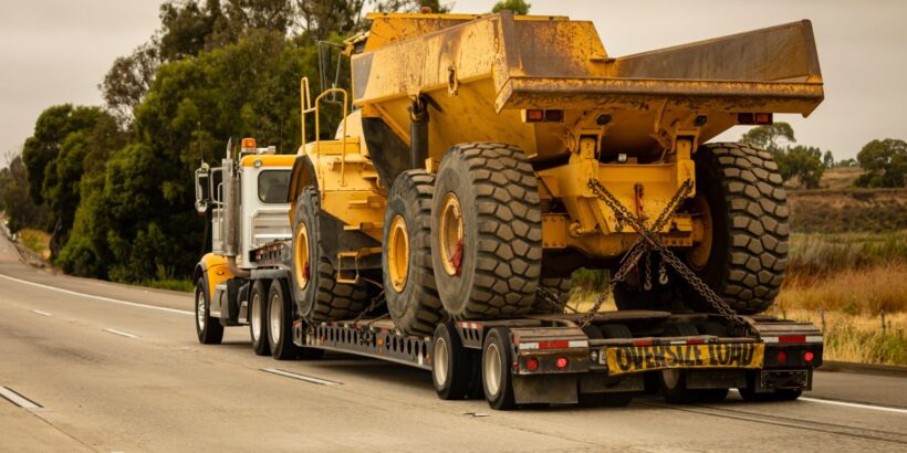 A dirty dump truck is strapped to the back of a semi. The semi is hauling the equipment down a three-lane road.