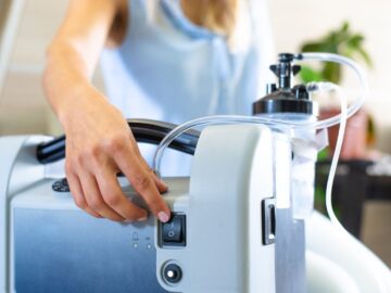 A close-up of a portable oxygen tank. Someone reaches around the device and touches the on-off switch.