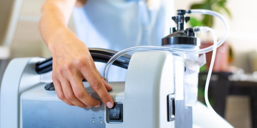 A close-up of a portable oxygen tank. Someone reaches around the device and touches the on-off switch.