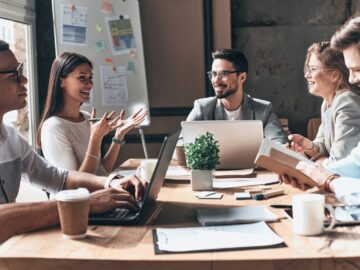 A group of professionals, with notebooks and laptops, gather around a table, smiling while having a meeting.