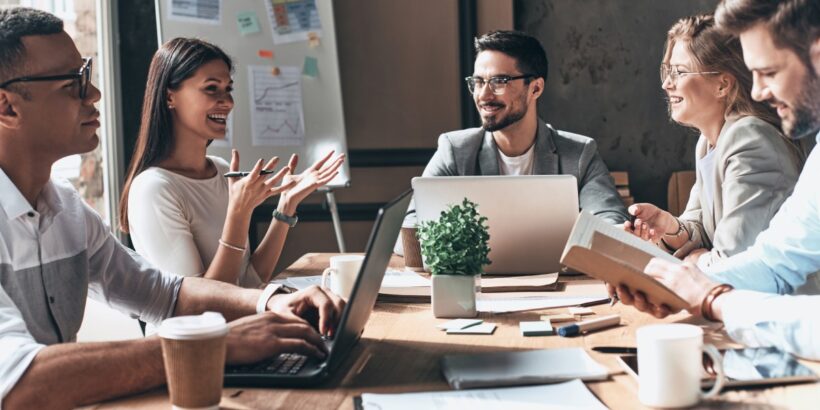 A group of professionals, with notebooks and laptops, gather around a table, smiling while having a meeting.