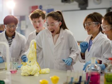A group of students standing around a bubbling science experiment. They look excited and are wearing safety gear.