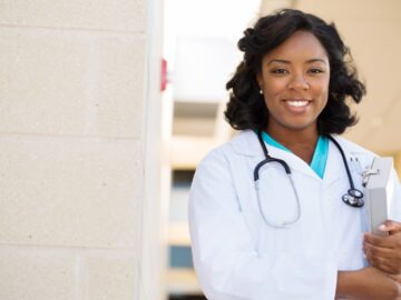 A smiling Black nurse practitioner stands outdoors in a white coat with a stethoscope and clipboard.