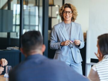 A manager smiling in an office. The manager stands between a whiteboard and a table full of employees.