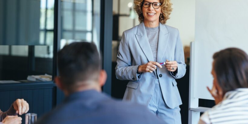 A manager smiling in an office. The manager stands between a whiteboard and a table full of employees.