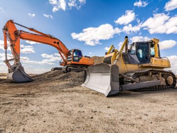 An orange excavator and a yellow bulldozer are positioned on a hilly, dirt-covered jobsite. A blue sky fills the background.