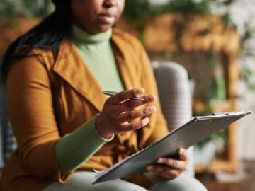A professional woman holds a clipboard in one hand and gestures with the other. Her eyes are not shown in the frame.