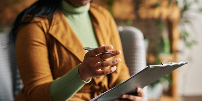 A professional woman holds a clipboard in one hand and gestures with the other. Her eyes are not shown in the frame.