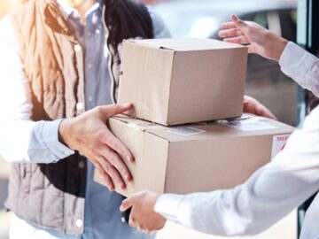 Two people exchange cardboard shipping boxes in a business setting, demonstrating package delivery and supply chain logistics.