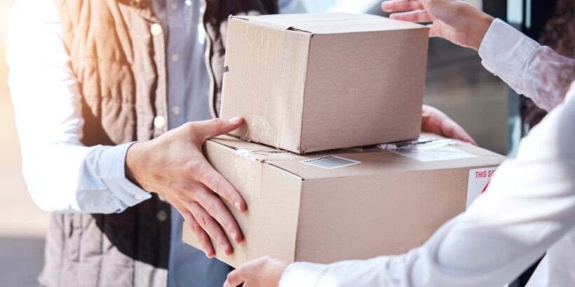 Two people exchange cardboard shipping boxes in a business setting, demonstrating package delivery and supply chain logistics.