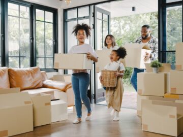 A Black family carrying boxes through the front door into a new home. There are boxes stacked against the wall.