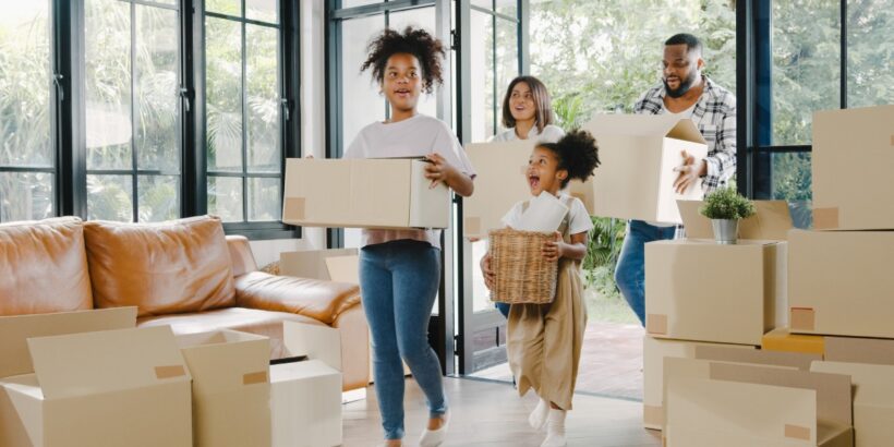 A Black family carrying boxes through the front door into a new home. There are boxes stacked against the wall.