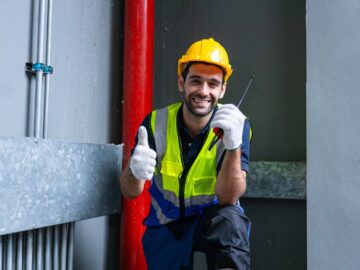 A man in safety gear holding a walkie-talkie and giving a thumbs up. He's kneeling down next to a red pipeline.