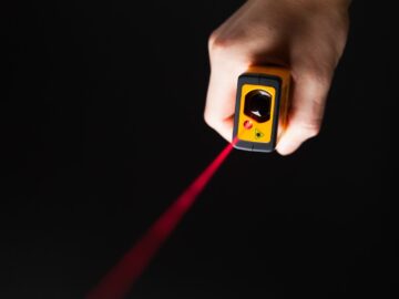 A hand holds a yellow laser-based gas detection device that emits a red beam against a dark background for safety monitoring.