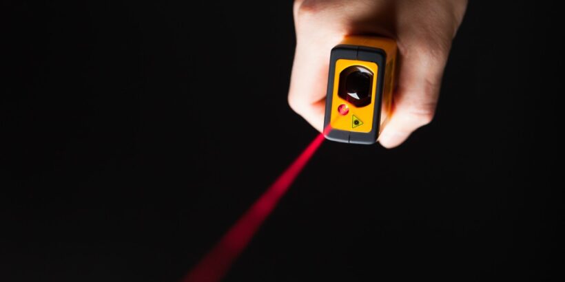 A hand holds a yellow laser-based gas detection device that emits a red beam against a dark background for safety monitoring.