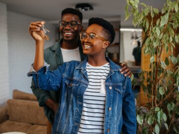 Two Black individuals, both wearing glasses and jackets, smile while holding keys as they stand indoors next to a plant.