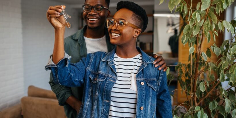 Two Black individuals, both wearing glasses and jackets, smile while holding keys as they stand indoors next to a plant.