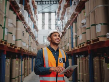 A warehouse worker in a hard hat and safety vest using a tablet in an aisle lined with tall shelves of boxed inventory.