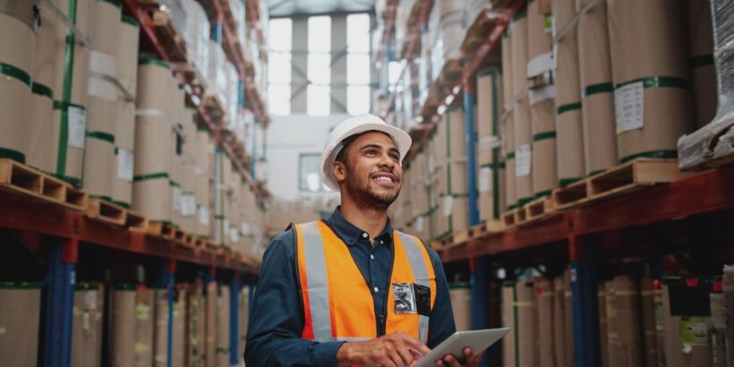 A warehouse worker in a hard hat and safety vest using a tablet in an aisle lined with tall shelves of boxed inventory.