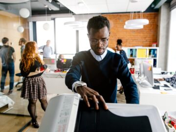 A young man places paper into a printer in a modern open office while coworkers collaborate in the background.