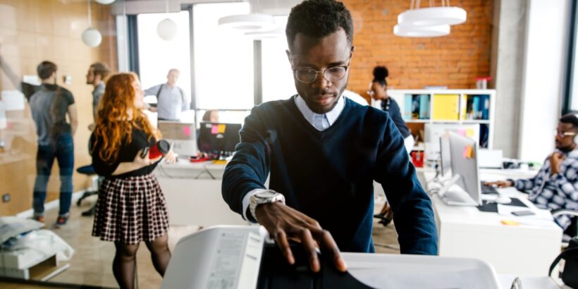 A young man places paper into a printer in a modern open office while coworkers collaborate in the background.