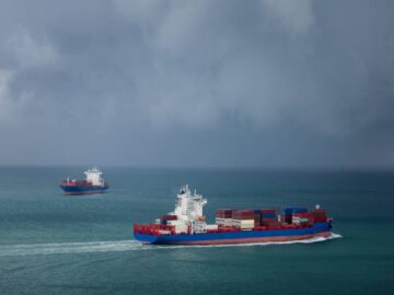 Two cargo container ships sailing past each other in the open ocean. The sky is filled with dark, gloomy clouds.